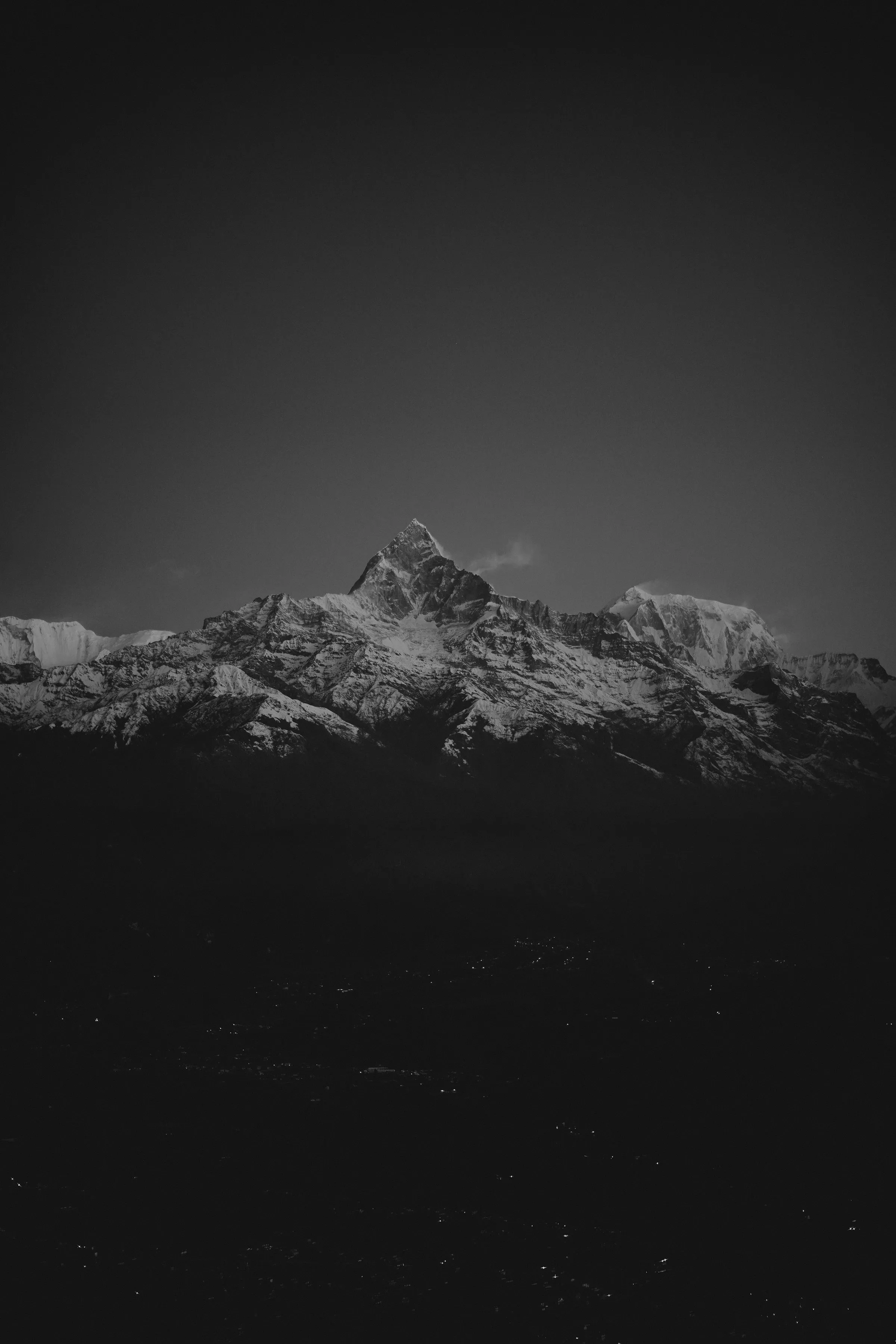 A moody black-and-white view of a Himalayan peak wreathed in cloud above a lit valley